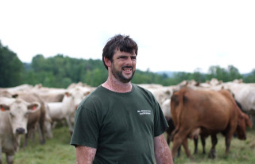 Brian Kemp is standing in a pasture wearing a dark green tee shirt. He looks off into the distance, a herd of brown and white cows are behind him.