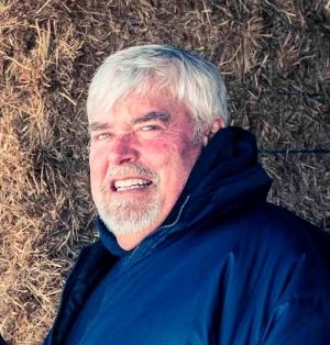 A head shot of Loren Wood smiling for the camera. Bales of hay are stacked behind him.