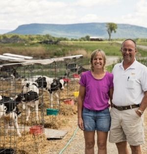Rob and Suzy Hunt pose beside the calf hutches at their farm. Several cute holstein calves are pictured. Snake Mountain is off in the distance.
