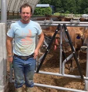 Caleb Smith is standing in the barn yard. A herd of jerseys are in the pen behind him. He is wearing jeans, boots caked in mud, and a light blue tee shirt.