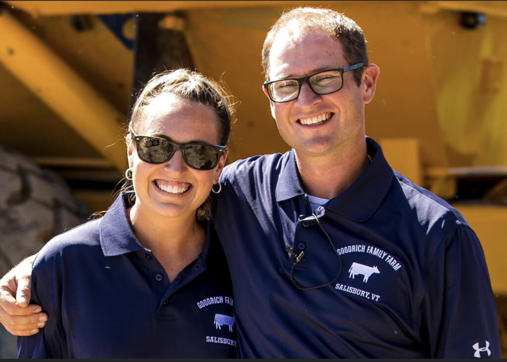 A photo of Danielle Goodrich and Chase Goodrich. Both are wearing navy blue polo shirts emblazoned with their farm's cow logo. They are smiling, and Chase has his arm wrapped around his sister's shoulder.