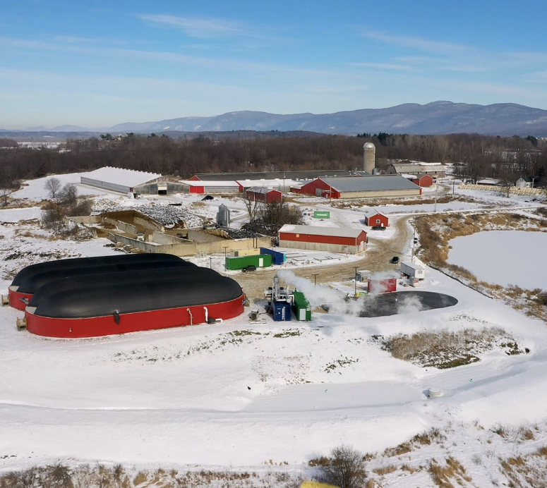 An aerial view of Goodrich Family Farm. The red barns are in the background. The red digester is in the foreground. The ground is covered in snow. Blue skies over the Green Mountains are in the distance.